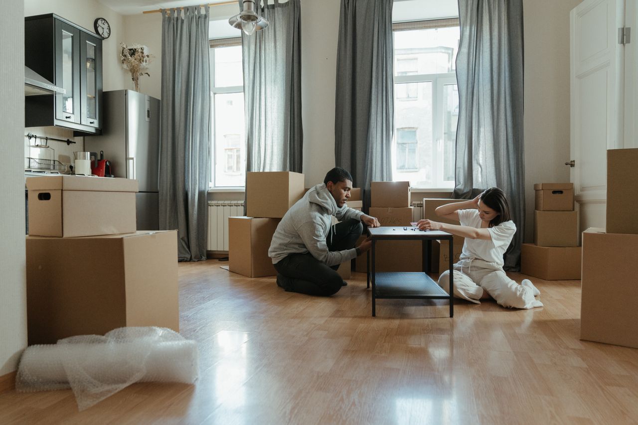 A young couple putting furniture together in their new apartment, with cardboard boxes all around them.